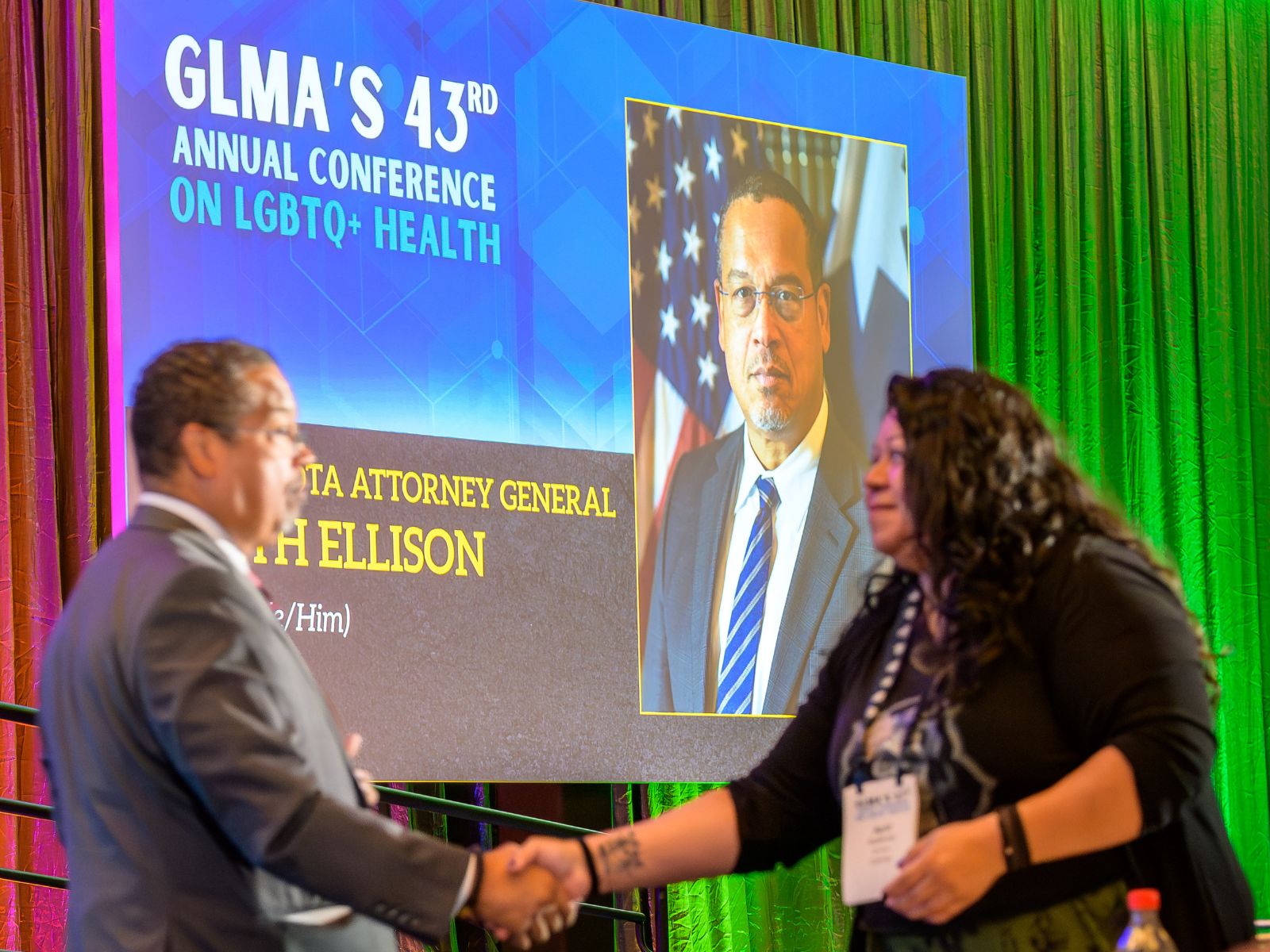 Presenter and attendee shaking hands in front of an LED screen featuring conference session information.